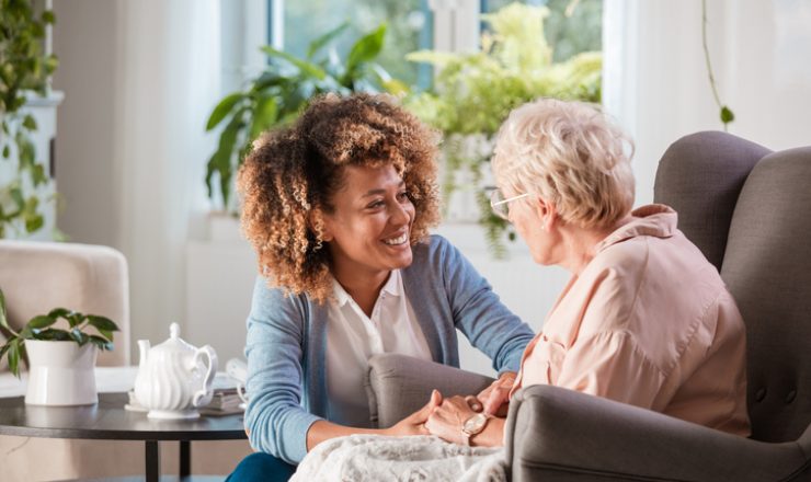 Female home caregiver talking with senior woman, sitting in living room and listening to her carefully.